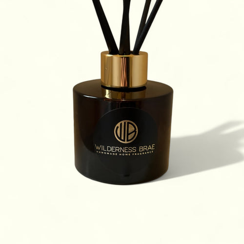 A black amber glass reed diffuser with a gold cap labeled 'Wilderness Brae' and white fibre reeds.