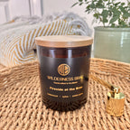 Fireside at the Brae soy candle with wooden lid on and Wilderness Brae, Handcrafted in Scotland black & gold label on front.  Amber glass jar and a cosy indoor background.