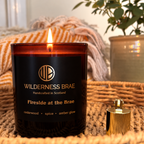 Fireside at the Brae lit soy candle labeled Wilderness Brae, Handcrafted in Scotland shown on a woven mat. A cozy blanket and potted plant are softly blurred in the background.