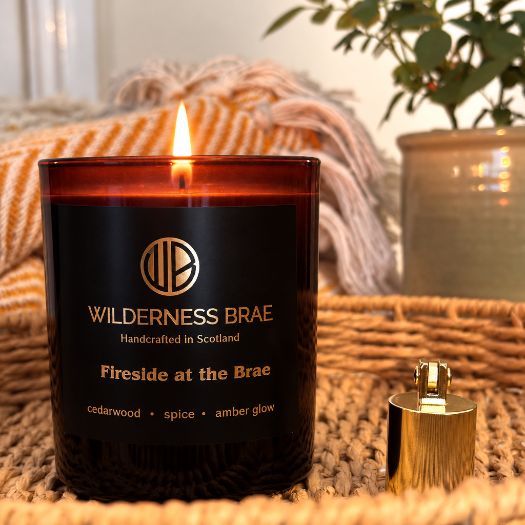 An amber glass candle labeled Wilderness Brae 'Fireside at the Brae' with a lit flame, shown on a woven mat with its gold-toned lid placed beside it. A cozy blanket and potted plant are softly blurred in the background.