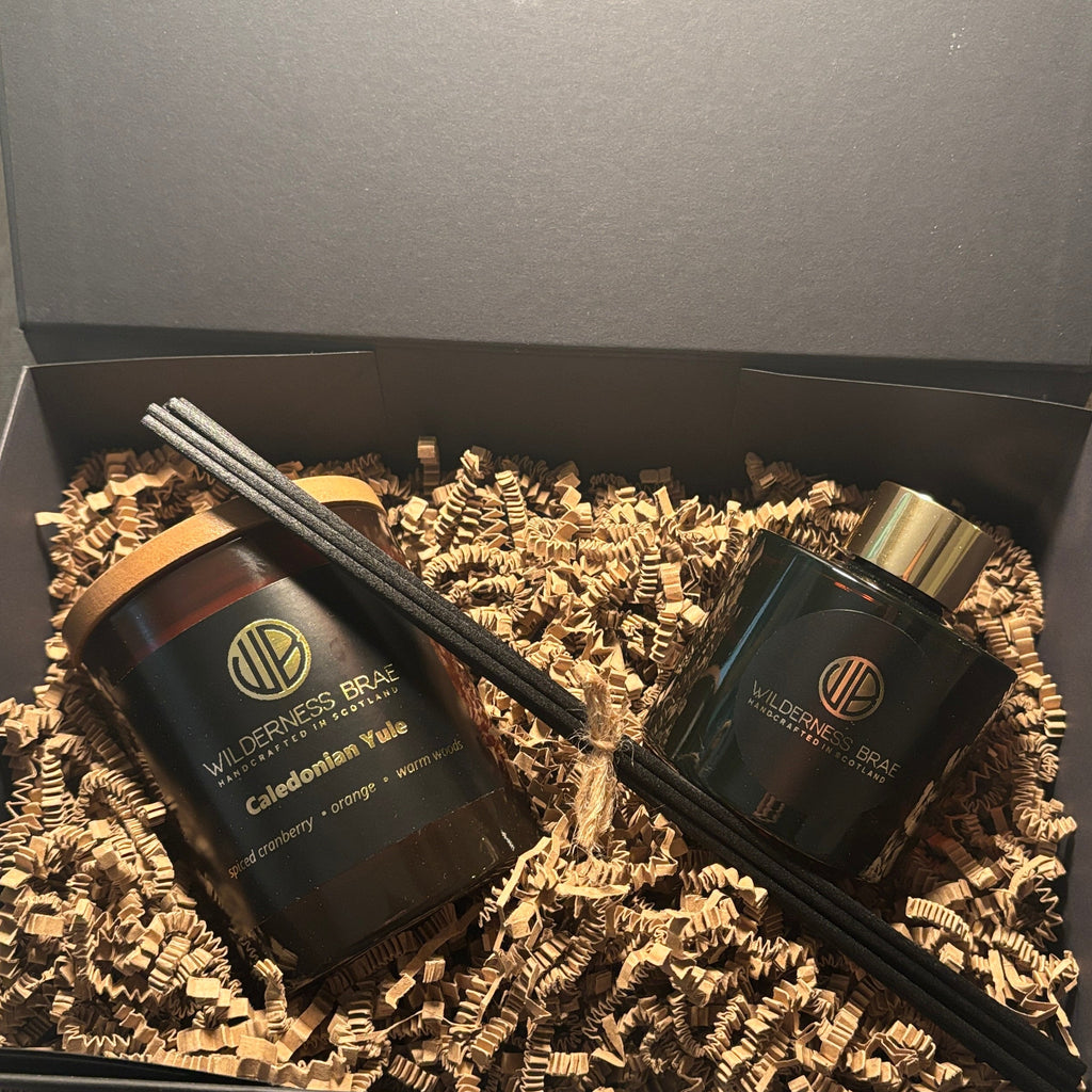 A gift set in an open dark gray box filled with brown crinkle paper, containing two black reed diffuser bottles with gold and white labels and several black reeds.