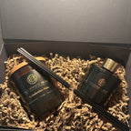A gift set in an open dark gray box filled with brown crinkle paper, containing two black reed diffuser bottles with gold and white labels and several black reeds.