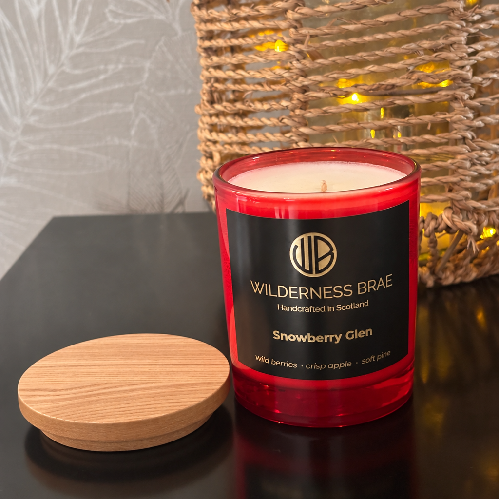 Snowberry Glen soy candle, with black and gold label reading Wilderness Brae, Handcrafted in Scotland.  Red glass jar candle & wooded lid resting beside it on a dark tabletop. A woven basket with warm string lights is blurred in the background.