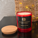 Snowberry Glen soy candle, with black and gold label reading Wilderness Brae, Handcrafted in Scotland.  Red glass jar candle & wooded lid resting beside it on a dark tabletop. A woven basket with warm string lights is blurred in the background.