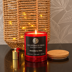 A red glass scented candle labeled Wilderness Braé Snowberry Glen, lit with a visible flame, on a dark reflective surface in front of a woven basket with warm string lights.