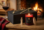 Wilderness Brae Still & Bright Box with  'Snowberry Glen' scented candle in a red glass jar with its wooden lid, a small match bottle, and brass candle tongs on a black gift box against a cosy fire in the background