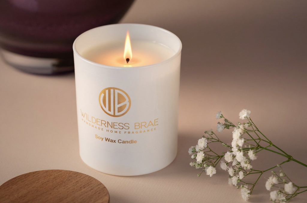 White candle with 'Wilderness Brae' branding on a beige surface with white flowers.