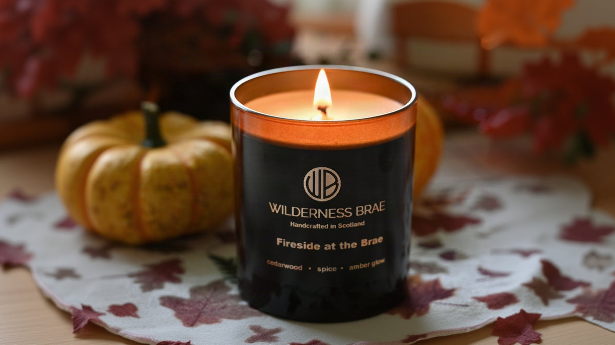 A lit Wilderness Brae candle in a black glass jar with a warm amber-orange wax surface, placed on a floral tablecloth with pumpkins and autumn decor blurred in the background.