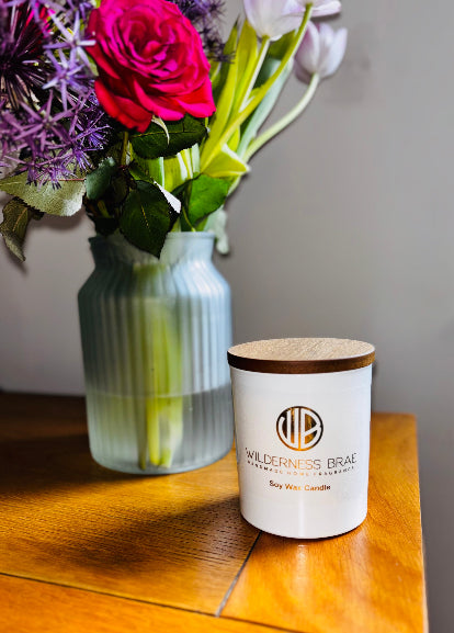 White Candle with wooden lid on a wooden surface next to a vase of flowers