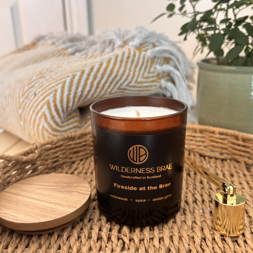 Fireside at the Brae soy candle on woven tray with lid off. Amber glass jar with a black label reading 'WILDERNESS BRAE Handcrafted in Scotland' featuring a striped blanket and a potted plant.