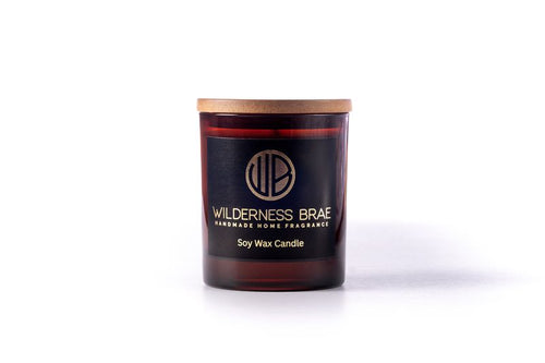 Caledonian Yule soy candle handcrafted by Wilderness Brae. Amber jar with black & gold logo and wooden lid on white background 

