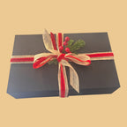 Wilderness Brae Still & Bright Candle Gift box - a black gift box tied with red and beige ribbons, decorated with a small green pine sprig and red berries, on a solid beige background.