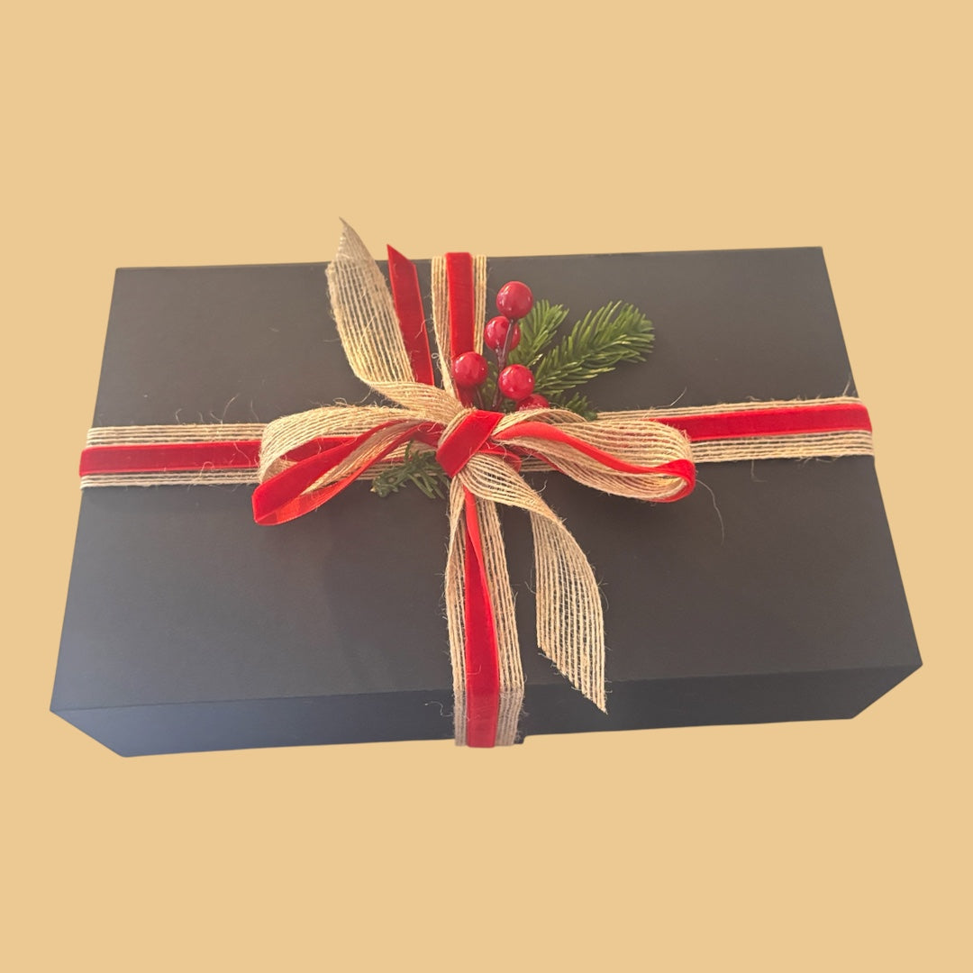 Wilderness Brae Still & Bright Candle Gift box - a black gift box tied with red and beige ribbons, decorated with a small green pine sprig and red berries, on a solid beige background.